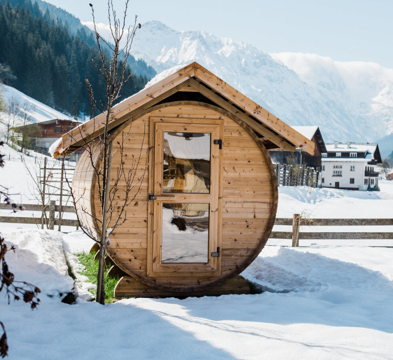 Fasssauna mit Tür und Türfenster im tiefen Schnee vor verschneitem Bergpanorama