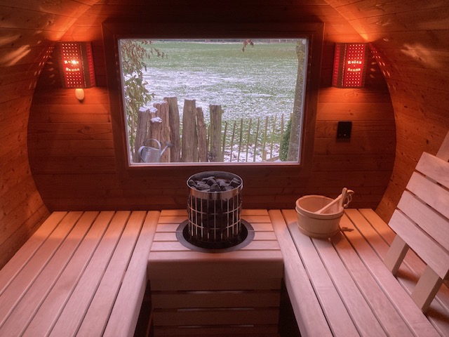 Warmer Innenraum einer Fasssauna mit Saunaofen, Aufgusskübel und Fenster zum verschneiten Garten